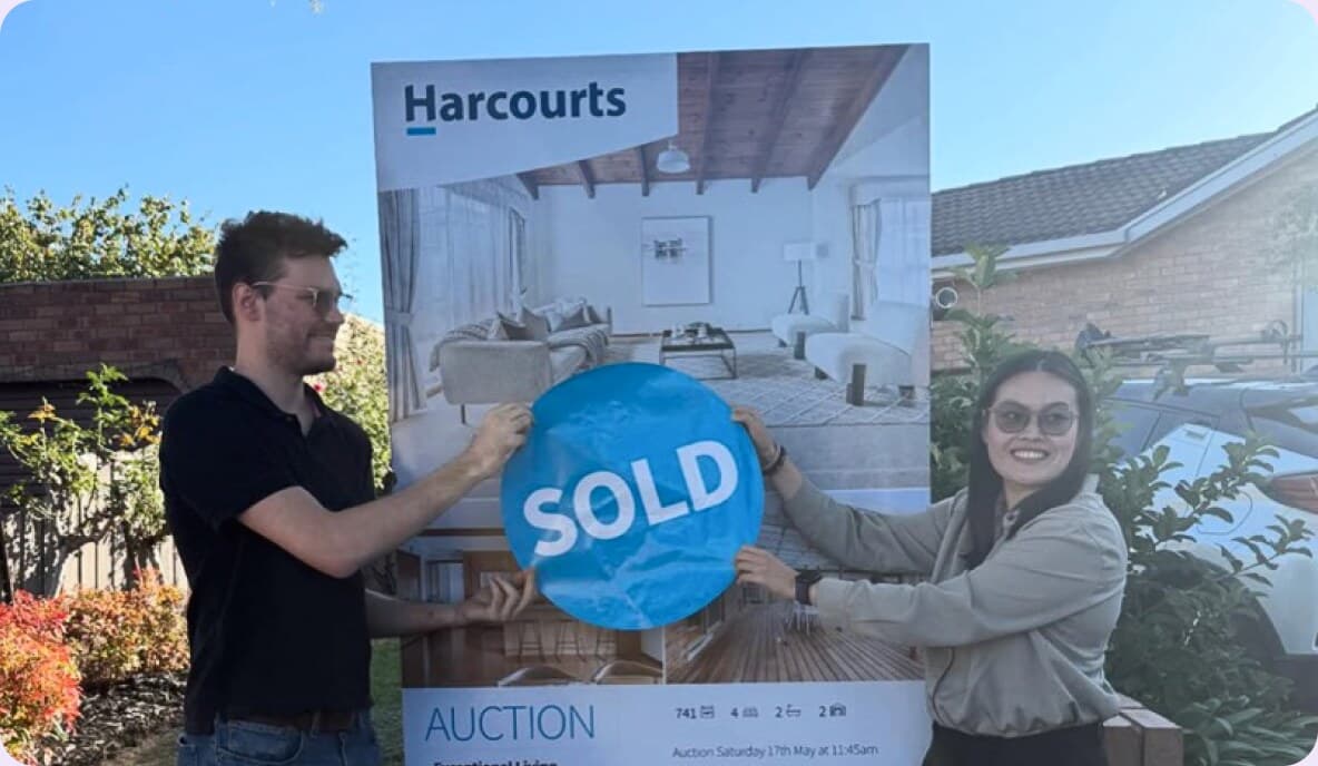 Buyers holding a SOLD sticker over an auction board after buying their home.